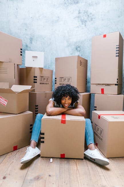 A young woman with curly black hair and a bright smile is sitting on the wooden floor inside a room, surrounded by numerous sealed cardboard moving boxes of various sizes. Some boxes have red and blac