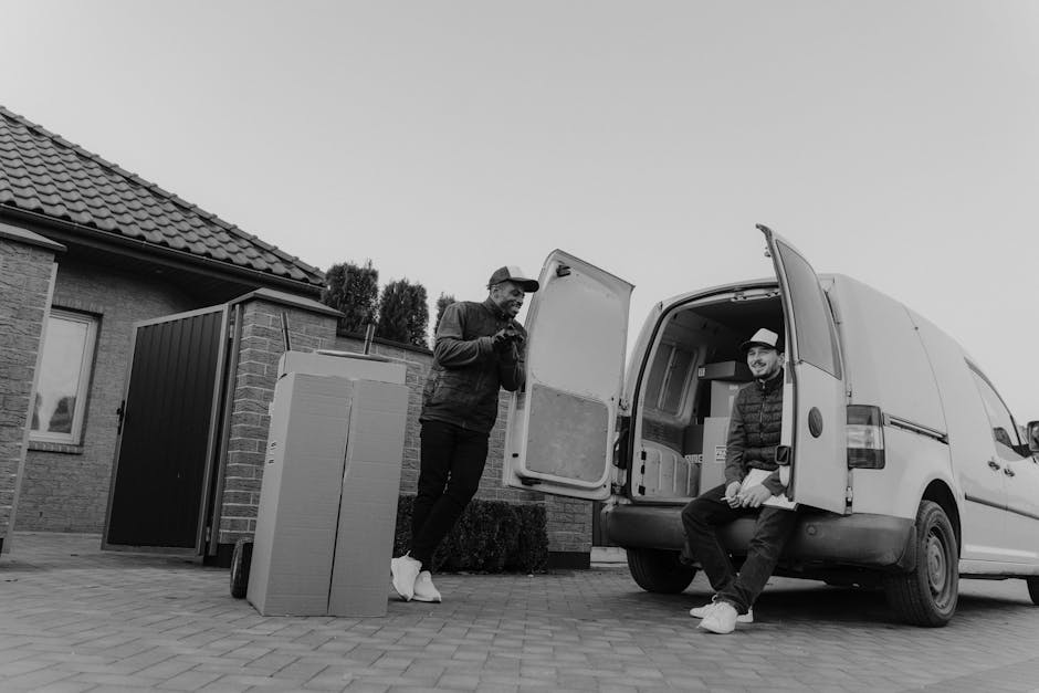 Two movers from Man and Van Chessington are engaged in a home relocation process, loading a large cardboard box wrapped in plastic transfer film into the back of a white cargo van parked on a paved dr