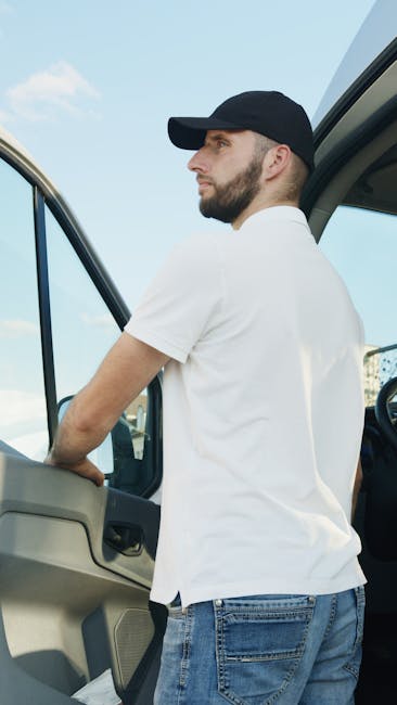 A professional man with a beard, wearing a black cap and white t-shirt, is standing outside a large moving van during daytime, with the driver’s side door open. He is preparing for a home relocation, with various cardboard boxes of different sizes and shapes, some wrapped in plastic or fabric, neatly stacked inside the vehicle. The boxes are being loaded or unloaded onto a trolley or hand truck on the pavement, indicating organized packing and furniture transport in progress. The scene is set outdoors against a light blue sky with some clouds, and the van is parked near the doorway of a residence. This image, representing loading and packing activities, is associated with house removals and the moving process, aligning with services offered by Man and Van Chessington, as referenced on the webpage for KT9 same-day removal rates for Chessington.
