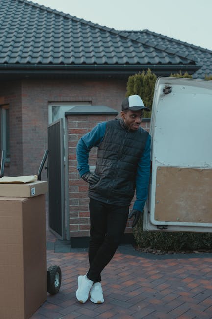 A man wearing a grey and white cap, black gloves, a dark padded vest over a blue long-sleeved shirt, and black trousers is standing next to an open white and brown removal van parked outside a residential property. He is leaning against the rear door of the van, which is partly open, revealing the empty interior used for transporting household goods. The van is positioned on a paved driveway with a brick house in the background, featuring a tiled roof, a window, and an open garden gate. Several cardboard boxes, some wrapped in plastic or packing materials, are placed nearby on the driveway, ready for a home relocation. The setting appears to be early morning or late afternoon, with soft natural light illuminating the scene, capturing the loading process associated with professional removals by Man and Van Chessington during furniture transport and moving operations.