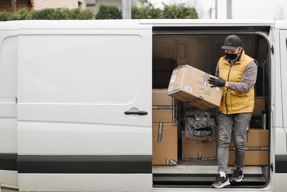 A professional man with a beard, wearing a black cap and white t-shirt, is standing outside a large moving van during daytime, with the driver’s side door open. He is preparing for a home relocation, with various cardboard boxes of different sizes and shapes, some wrapped in plastic or fabric, neatly stacked inside the vehicle. The boxes are being loaded or unloaded onto a trolley or hand truck on the pavement, indicating organized packing and furniture transport in progress. The scene is set outdoors against a light blue sky with some clouds, and the van is parked near the doorway of a residence. This image, representing loading and packing activities, is associated with house removals and the moving process, aligning with services offered by Man and Van Chessington, as referenced on the webpage for KT9 same-day removal rates for Chessington.