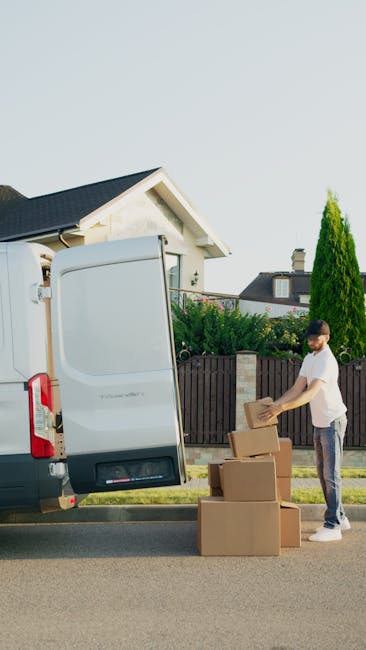 A man wearing a white t-shirt and jeans is standing on the pavement next to a white moving van with its rear doors open, preparing to load or unload cardboard boxes during a home relocation. He is holding a cardboard box, while several other boxes are stacked on the ground nearby. The scene takes place in a residential area with a house visible in the background, featuring a dark tiled roof, white walls, and a garden with green bushes and a tall, narrow tree. The moving van, equipped with a tail lift, is positioned close to the property, suggesting a furniture transport or packing and moving process managed by Man and Van Chessington. The environment is well-lit with natural daylight, and the overall scene depicts typical logistics involved in house removals within the Chessington area.