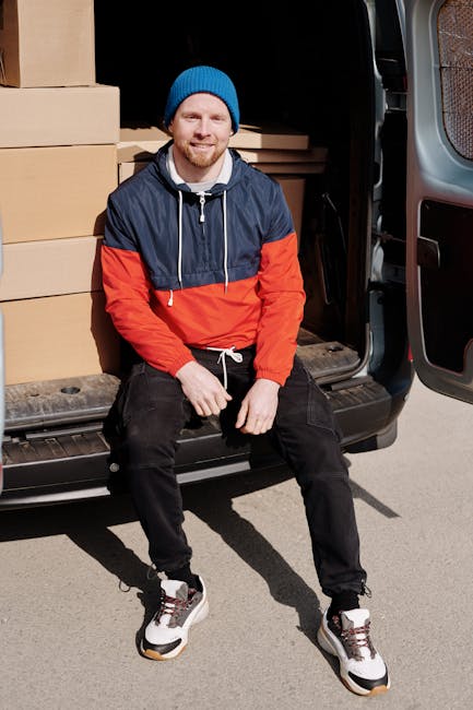 Two men wearing dark jackets and gloves are engaged in a home relocation task, lifting and carrying a large cardboard box from a white moving van parked on a paved driveway. The van's rear doors are open, revealing more boxes and packing materials inside. One man is standing inside the van, helping to guide the box, while the other is outside, lifting and maneuvering it onto a black hand truck with two wheels, positioned nearby. The driveway is bordered by a brick wall and a small hedge, with a house featuring a tiled roof visible in the background. During the loading process, the scene is illuminated by natural daylight under an overcast sky, illustrating the packing and furniture transport aspects involved in house removals, with Man and Van Chessington providing the logistical support for such moving services.
